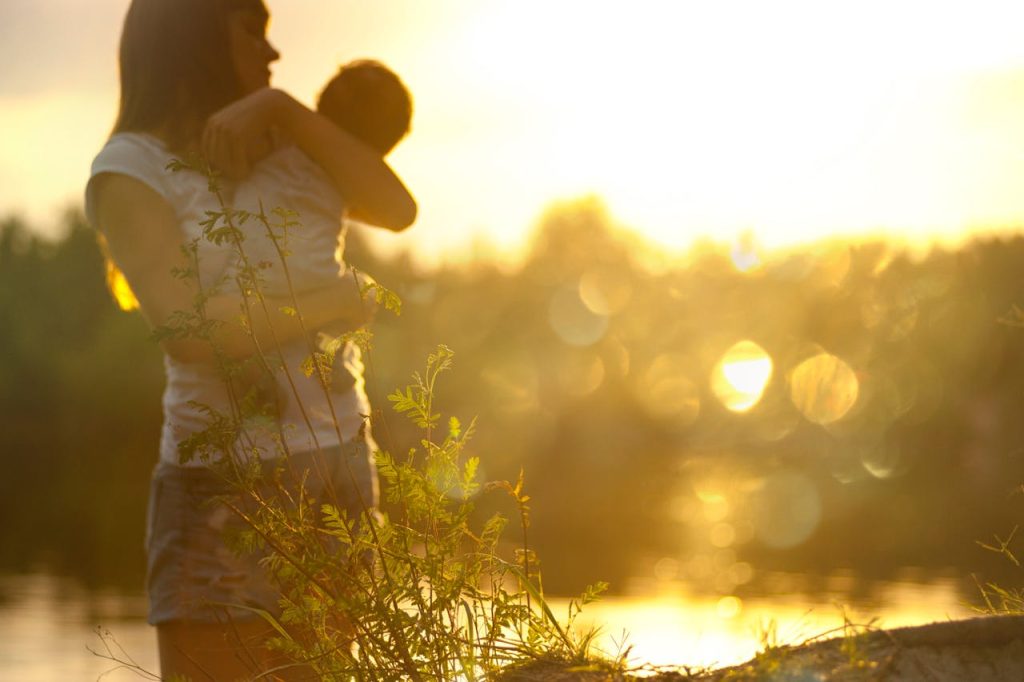 A serene moment of a mother holding her baby at sunset by a lake.